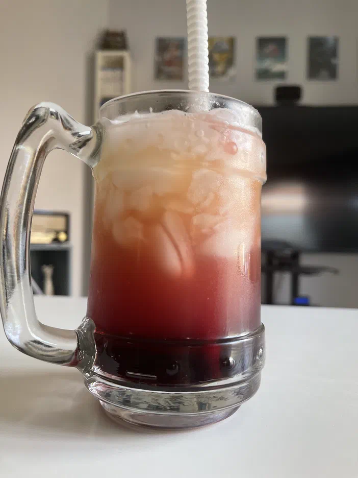 Un verre à bière en forme de mug rempli d'un cocktail rouge et blanc avec des glaçons,&nbsp;une paille blanche et un fond flou montrant un intérieur.